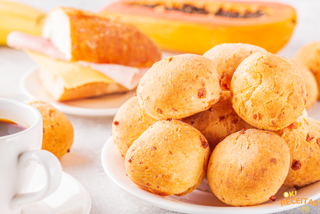 Pães de queijo dispostos sobre um prato branco, com outros alimentos desfocados, ao fundo.