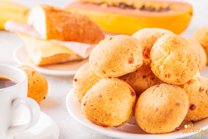 Pães de queijo dispostos sobre um prato branco, com outros alimentos desfocados, ao fundo.